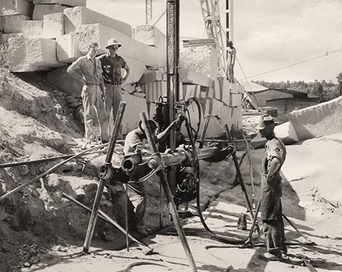 For centuries, man quarried stone using a hand-held bit which was struck with heavy hammers. With the advent of pneumatic, or air-powered tools, the channel bar drill (above) was developed which greatly sped up the broaching or cracking loose of large sections of stone. The channel bar was basically a huge jackhammer with a vertical drill support. Drill bits as long as 12 feet moved horizontally via a transversing beam mounted on A-Frame legs. The holes were drilled from four inches to six inches apart the entire length and width of the section to be freed. Then the drill was replaced by a broaching bit or core cutter with teeth and the granite between the drill holes was removed. Loosened vertically on all sides, the block was freed on the bottom by drilling horizontal “lift” holes. The holes were loaded with explosives which caused horizontal lifting when detonated. The large section was then split into smaller blocks by hand-held jackhammers. 