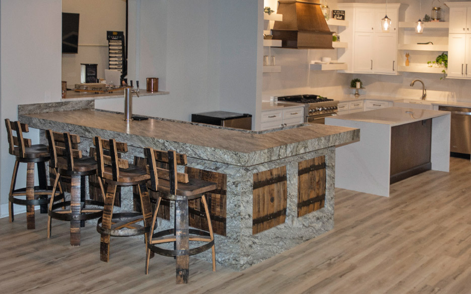 A fully-functional display kitchen in the Artistic Granite and Marble showroom features a working tap and appliances, theme-decorated with recycled bourbon casks repurposed as stools, light fixtures, and insets set in a granite bar. 